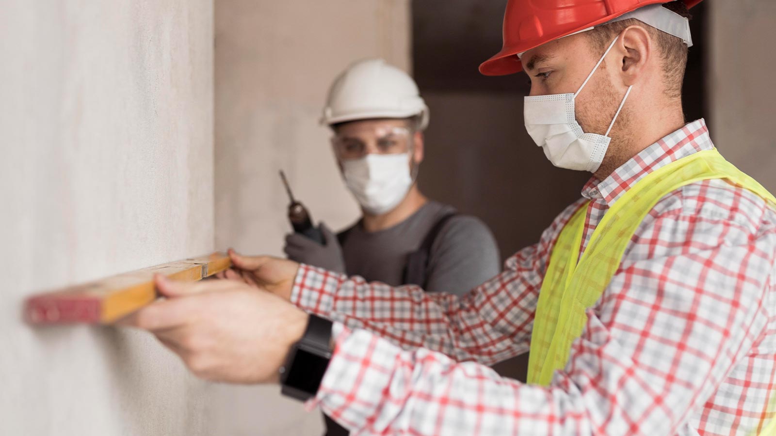 Zwei Handwerker mit Schutzmaske und Helm prüfen mit der Wasserwaage eine Wand – Arbeitsalltag bei der Sanitec GmbH